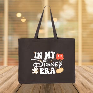 Puede incluir: Bolso tote negro con el texto blanco "IN MY Disney ERA", con gr&aacute;ficos tem&aacute;ticos de Mickey Mouse. El bolso tiene dos asas y est&aacute; hecho de una tela resistente. El fondo est&aacute; borroso, con una superficie de madera en primer plano.