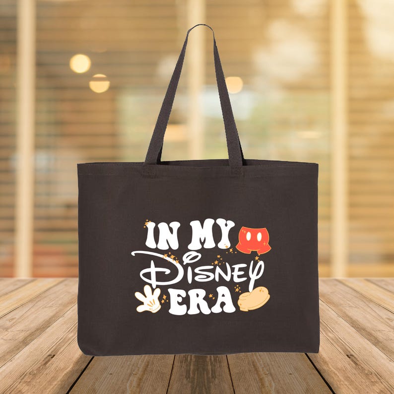 Puede incluir: Bolso tote negro con el texto blanco "IN MY Disney ERA", con gr&aacute;ficos tem&aacute;ticos de Mickey Mouse. El bolso tiene dos asas y est&aacute; hecho de una tela resistente. El fondo est&aacute; borroso, con una superficie de madera en primer plano.