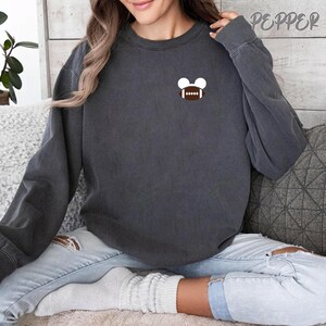 May include: Dark gray sweatshirt with a small football and mouse ears graphic on the chest. The word "Pepper" is printed in white on the upper right side. The sweatshirt is worn with light blue jeans.