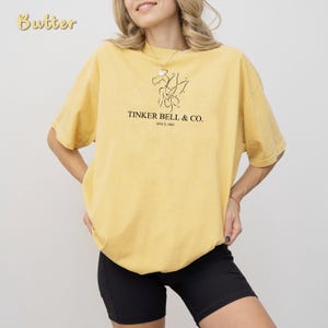 May include: Butter-colored, short-sleeved t-shirt with a crew neck. The shirt features a black graphic of a fairy and the text "TINKER BELL & CO. SINCE 1964". The model is wearing black shorts.