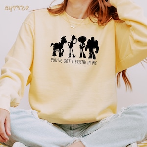 May include: A light yellow sweatshirt featuring a black graphic of four cartoon characters and the text "YOU'VE GOT A FRIEND IN ME". The sweatshirt has long sleeves and a crew neck.