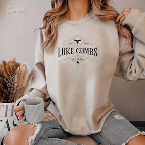 May include: A beige sweatshirt featuring the text "Luke Combs" in a black, vintage-style design. The design includes a bull skull and the words "Est 1990" and "The Legend". The sweatshirt is a neutral color.