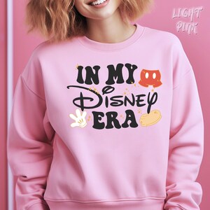 May include: Light pink sweatshirt with the text "IN MY Disney ERA" in black script and block letters. The design includes Mickey Mouse shorts, a white glove, and a yellow shoe. The sweatshirt is a crew neck.