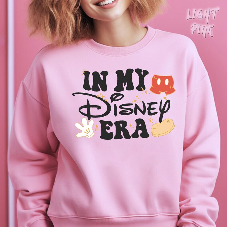 May include: Light pink sweatshirt with the text "IN MY Disney ERA" in black script and block letters. The design includes Mickey Mouse shorts, a white glove, and a yellow shoe. The sweatshirt is a crew neck.