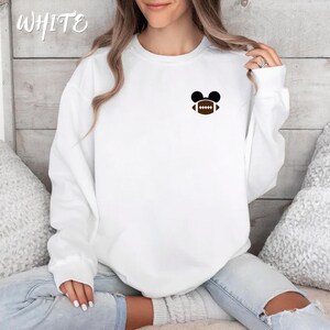 May include: White sweatshirt with a small graphic of a football inside Mickey Mouse ears. The word "WHITE" is written in cursive in the upper left corner. The sweatshirt is long-sleeved and has a crew neck.