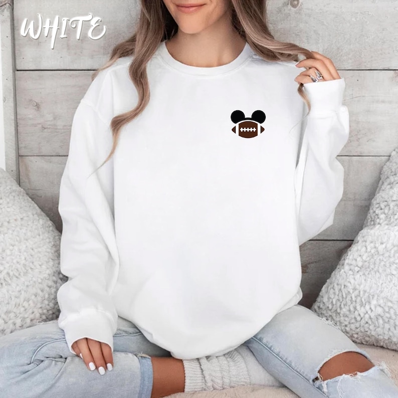 May include: White sweatshirt with a small graphic of a football inside Mickey Mouse ears. The word "WHITE" is written in cursive in the upper left corner. The sweatshirt is long-sleeved and has a crew neck.