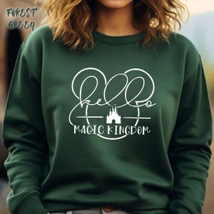 May include: Forest green sweatshirt with the text "Hello Magic Kingdom" in white script. The design includes a stylized castle and Mickey Mouse ears. The sweatshirt is a solid color and has a crew neck.