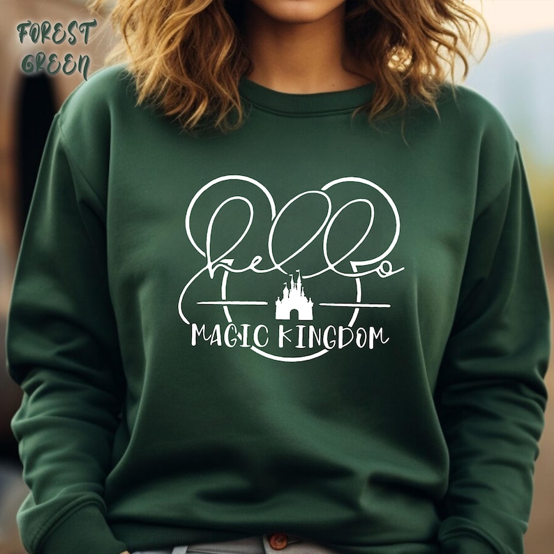 May include: Forest green sweatshirt with the text "Hello Magic Kingdom" in white script. The design includes a stylized castle and Mickey Mouse ears. The sweatshirt is a solid color and has a crew neck.