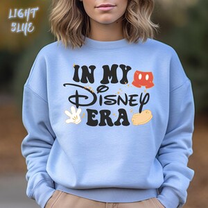May include: Light blue sweatshirt with the text "IN MY Disney ERA" in black, with Mickey Mouse-themed graphics in red, white, and yellow. The words "LIGHT BLUE" are in the upper left corner.