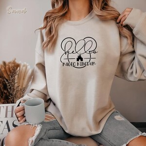 May include: Beige sweatshirt with the text "Hello Magic Kingdom" in black script and a castle graphic. The sweatshirt is a neutral color and has a crew neck. A light gray mug is held in the hand.