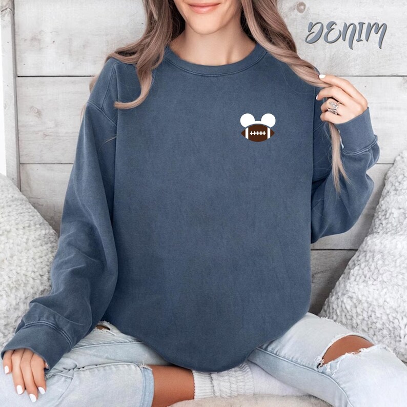 May include: Denim blue sweatshirt with a small football and Mickey Mouse ears design. The sweatshirt is a solid color and has a crew neck. The word "Denim" is written in cursive in the upper right corner.