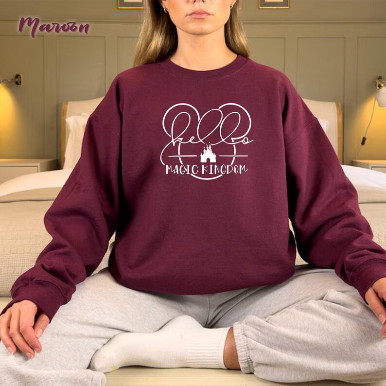 May include: A maroon sweatshirt with long sleeves. The front of the sweatshirt features the words "Hello Magic Kingdom" in white script and a castle graphic. The sweatshirt is a solid color and appears to be made of a soft material.