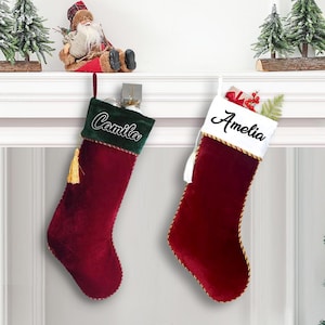May include: Two Christmas stockings, one with "Camila" embroidered on a green cuff and a gold tassel, the other with "Amelia" on a white cuff. Both are burgundy velvet with gold trim.