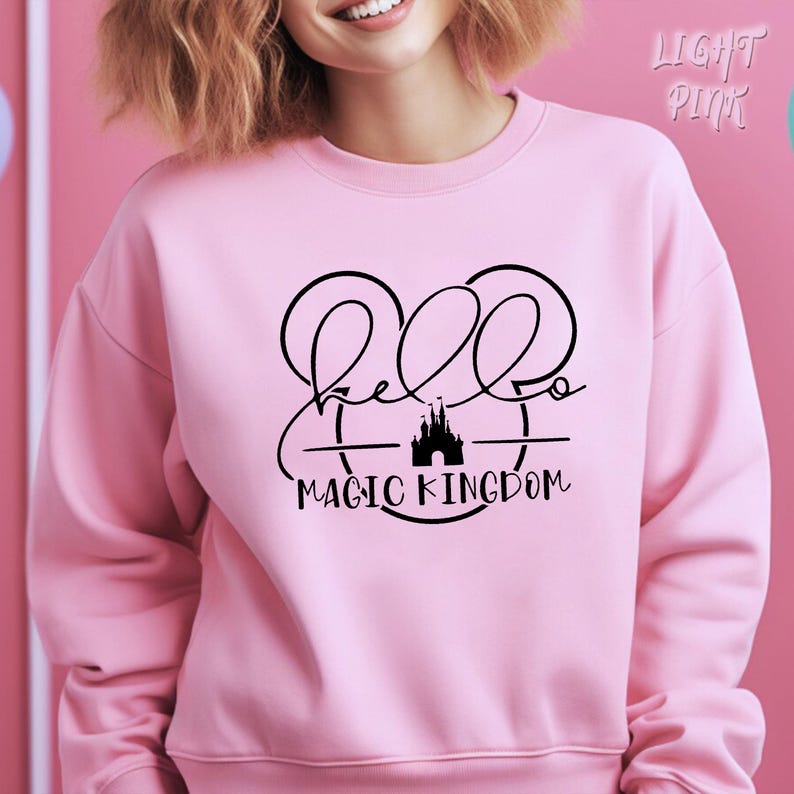 May include: A light pink sweatshirt with a black graphic that says "Hello Magic Kingdom". The graphic features Mickey Mouse ears and a castle silhouette. The sweatshirt is a crew neck style.