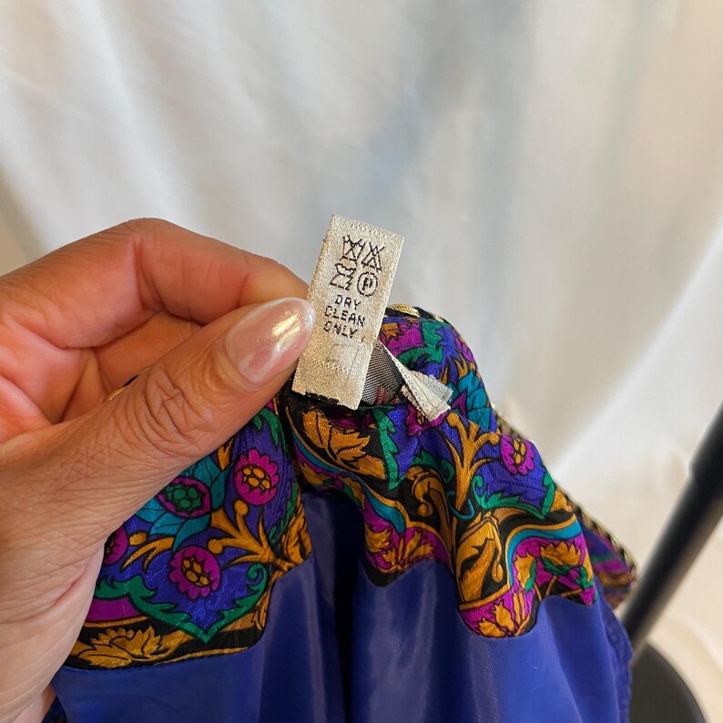 May include: Close-up of a garment with a vibrant, ornate border featuring gold, purple, and green floral patterns. The garment is a deep blue color. A tag reads "Dry Clean Only."