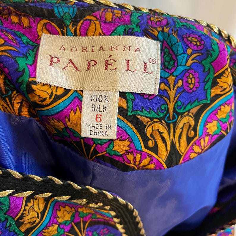 May include: Close-up of a colorful silk garment with a label that reads "ADRIANNA PAPELL". The garment features a vibrant floral pattern in shades of purple, blue, and gold. The label indicates the garment is 100% silk, size 6, and made in China.