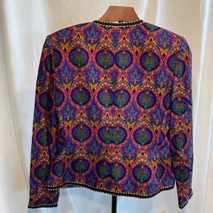 May include: A long-sleeved jacket with a vibrant, ornate pattern in shades of purple, blue, gold, and green. The jacket has a black trim with a white and gold rope-like design along the collar and hem. The jacket is displayed on a wooden hanger.
