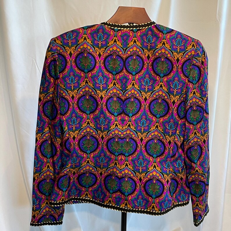 May include: A long-sleeved jacket with a vibrant, ornate pattern in shades of purple, blue, gold, and green. The jacket has a black trim with a white and gold rope-like design along the collar and hem. The jacket is displayed on a wooden hanger.