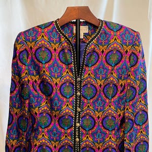 May include: A colorful jacket with a vibrant paisley pattern in shades of purple, blue, yellow, and pink. The jacket has a black trim with white stitching and gold buttons down the front. The jacket is hanging on a wooden hanger.