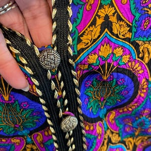 May include: Close-up of a colorful patterned fabric with a black braided trim and gold accents. The fabric features a vibrant mix of purple, blue, and gold floral designs. Silver-colored buttons secure the trim, adding a touch of elegance to the textile.