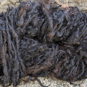 Puede incluir: Un ovillo de lana de color marrón oscuro y negro. La lana es muy rizada y tiene un aspecto natural y rústico.