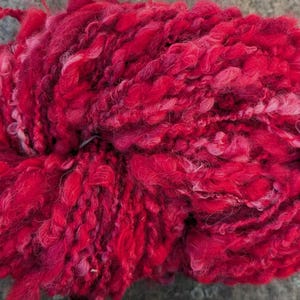 May include: A close-up of a vibrant red yarn ball. The yarn has a thick, textured appearance with varying shades of red and pink. The fibers are loosely spun, creating a soft, fluffy texture. Ideal for knitting and crochet projects.