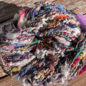 May include: A multi-colored skein of art yarn featuring a variety of textures, including fuzzy, metallic, and thick-spun strands in shades of blue, pink, orange, yellow, and white. A purple label is attached with visible text.