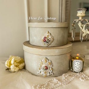 May include: Two stacked round light gray boxes with lids. The top box features a heart-shaped floral emblem, and the bottom box has an ornate rectangular floral plaque. The text Fleur La Vintage is visible above the boxes, alongside a candle and pearls.