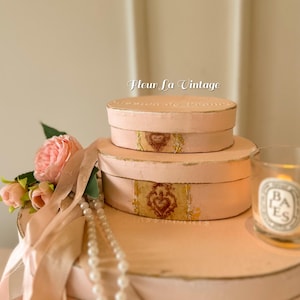 May include: Two stacked oval boxes in a soft pink hue with gold-painted edges. Each box features a central decorative label with a brown ornate emblem. The text Fleur La Vintage is visible above the boxes, and a string of pearls and pink flowers are placed alongside.