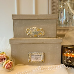 Op de afbeelding: Twee beige rechthoekige dozen, op elkaar gestapeld. Elke doos heeft een sierlijk, goudkleurig label. De onderste doos heeft de tekst "QUE DE PASSEMENT HAUTE-NOUVEAUTES. PARIS." Een kaars en rozen staan op de achtergrond.