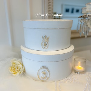 May include: Two white, oval-shaped boxes stacked on top of each other. The boxes have gold trim and a gold emblem on the front. The text "Fleur La Vintage" is written in cursive on the top of the image.