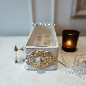 May include: A white rectangular decorative box with gold detailing and the text "Fleur La Vintage". The box features an ornate gold design on the front. A lit candle in a black glass and a pearl necklace are nearby.