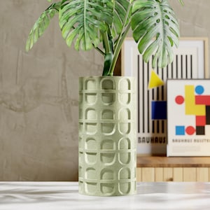 May include: A light green cylindrical planter with a repeating geometric pattern, holding a large-leafed plant. The planter is approximately 12 inches tall. Decorative framed art pieces are in the background.