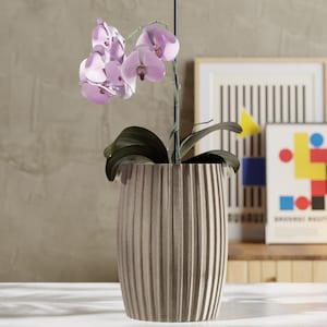 May include: A decorative beige ribbed planter holds a blooming orchid with light purple flowers and dark green leaves. The planter sits on a white surface, with framed art in the background.