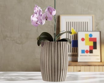 May include: A decorative beige ribbed planter holds a blooming orchid with light purple flowers and dark green leaves. The planter sits on a white surface, with framed art in the background.