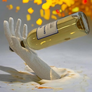 May include: A white wine bottle held by a white, textured hand-shaped sculpture. The bottle has a label with the word "GRANGE" and is filled with a light yellow liquid. The sculpture appears to be emerging from a white, textured surface.