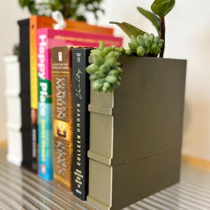 May include: A decorative bookend with a gold finish, holding several books and small plants. The books have colorful spines, including titles like "Happy Place" and "Verity". The arrangement sits on a clear, ridged surface.