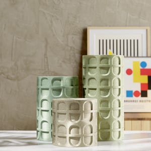 May include: Four cylindrical vases in varying sizes and shades of green and beige. Each vase features a repeating arched pattern. The vases are arranged in front of framed artwork, creating a modern, minimalist aesthetic.