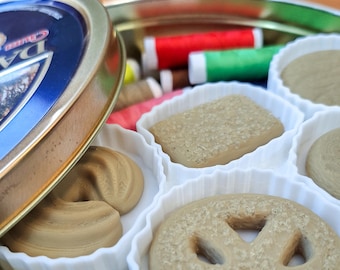 May include: A vintage sewing kit tin with a blue lid, partially open to reveal spools of thread and various shaped, light brown soaps in white, fluted containers. The soaps include a pretzel, a square, and a donut shape.