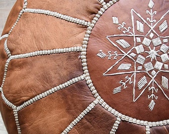May include: A brown leather pouf with white stitching in a geometric pattern. The pouf has a star-shaped design in the center.