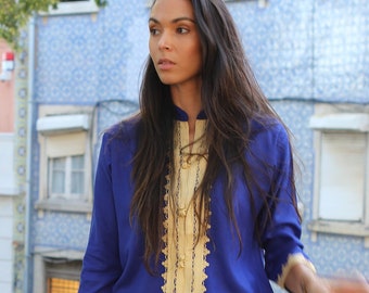 May include: A woman wearing a blue and gold embroidered shirt, white denim shorts, and white sandals. She is holding a straw hat and a woven bag.