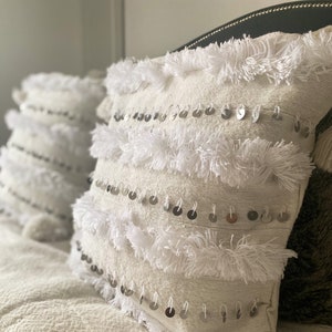 May include: Two white throw pillows with fringe and silver sequins on a cream-colored bedspread. The pillow on the right has a white pom-pom.