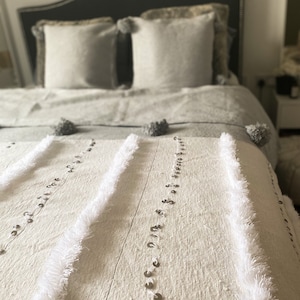 May include: A white throw blanket with a fringe border and silver sequins. The blanket is folded on a bed with gray pillows and a gray bedspread.