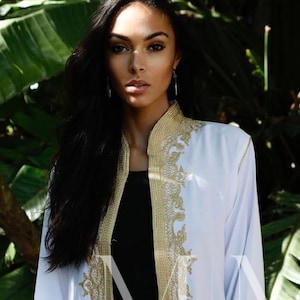 Könnte beinhalten: Eine weiße, langärmelige Robe mit goldener Stickerei. Die Robe ist vorne offen und zeigt darunter ein schwarzes Oberteil und eine blaue Jeans. Das Model steht in einem üppigen grünen Garten.