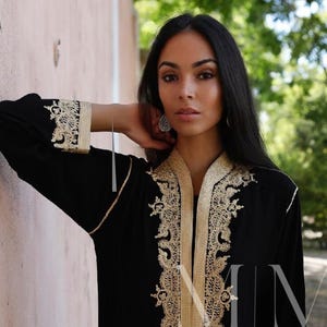 Puede incluir: Una túnica negra de manga larga con bordados dorados y el texto "Maison Marrakech" en la parte delantera.