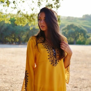 May include: A woman wearing a yellow tunic with black embroidery. The tunic has long sleeves and a V-neckline. The woman is standing in a field with green grass and trees in the background.