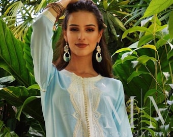 May include: A light blue kaftan with white embroidery and a white trim. The kaftan is worn by a person standing in front of a green leafy background. The text "MM Maison Marrakech" is visible in the image.