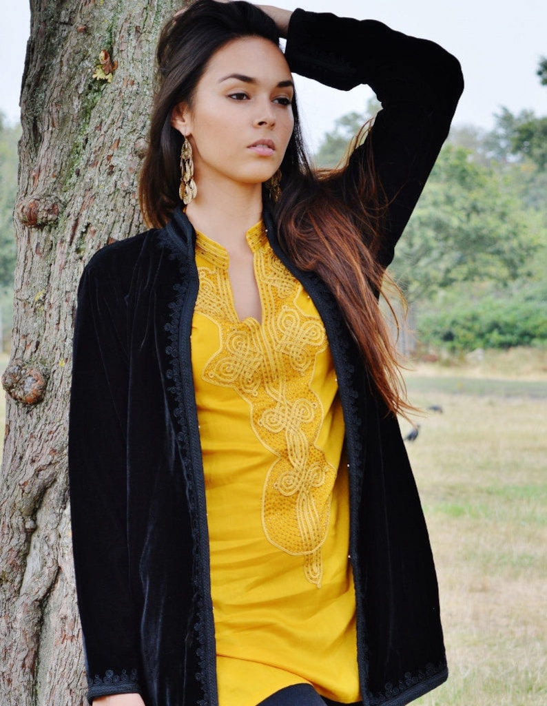 May include: A woman wearing a black velvet jacket with gold embroidery over a yellow top. She is standing next to a tree.