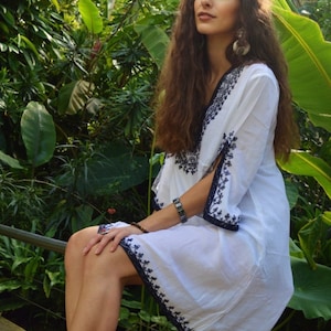 May include: A woman wearing a white dress with black embroidery sits in a lush green garden. The dress has a V-neckline and a flowing silhouette. She is wearing a bracelet on her wrist.