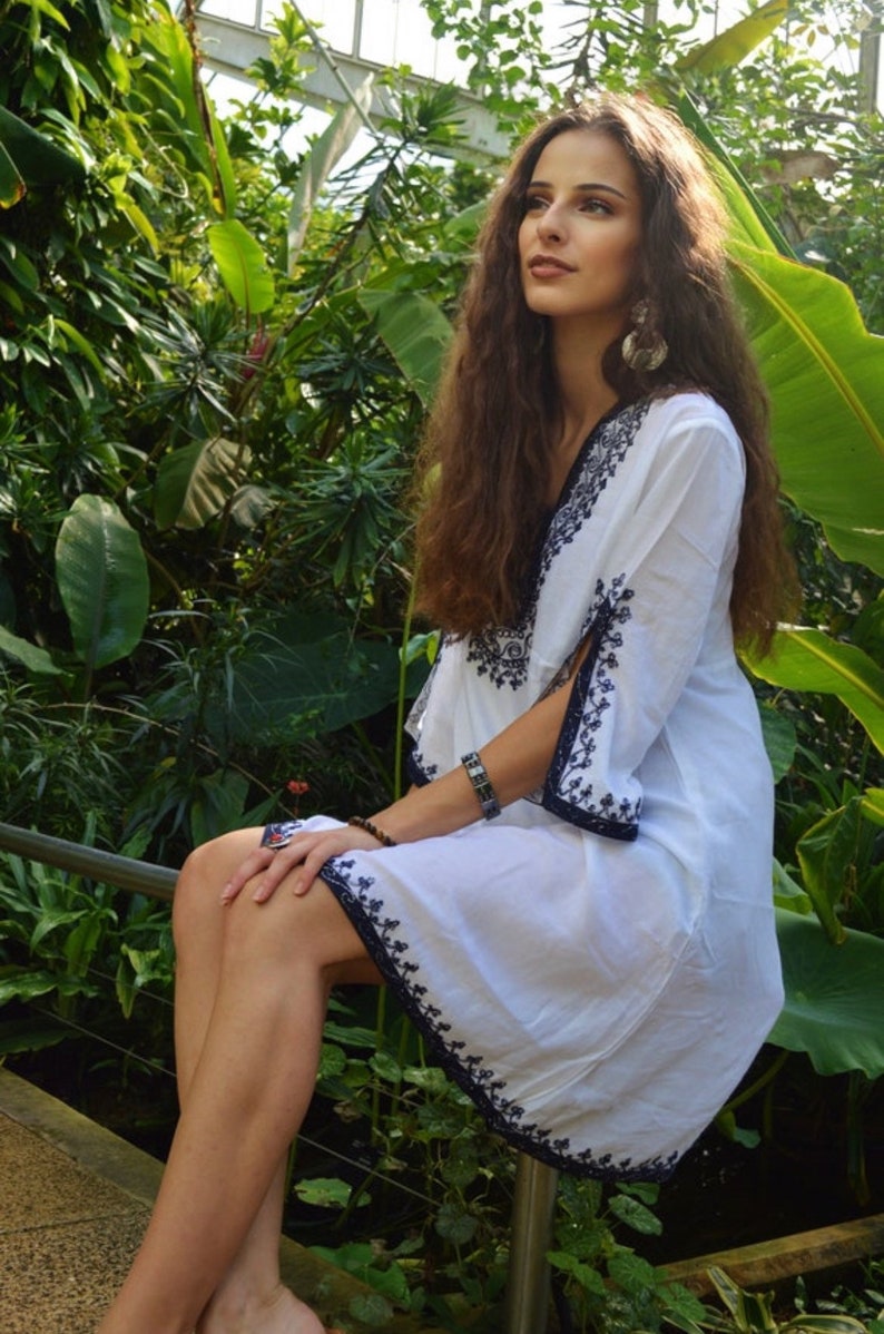 May include: A woman wearing a white dress with black embroidery sits in a lush green garden. The dress has a V-neckline and a flowing silhouette. She is wearing a bracelet on her wrist.